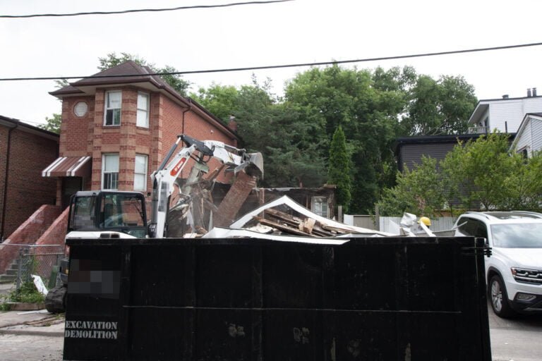 large demolition bin with in the background an excavator that is taking down a house chimney
