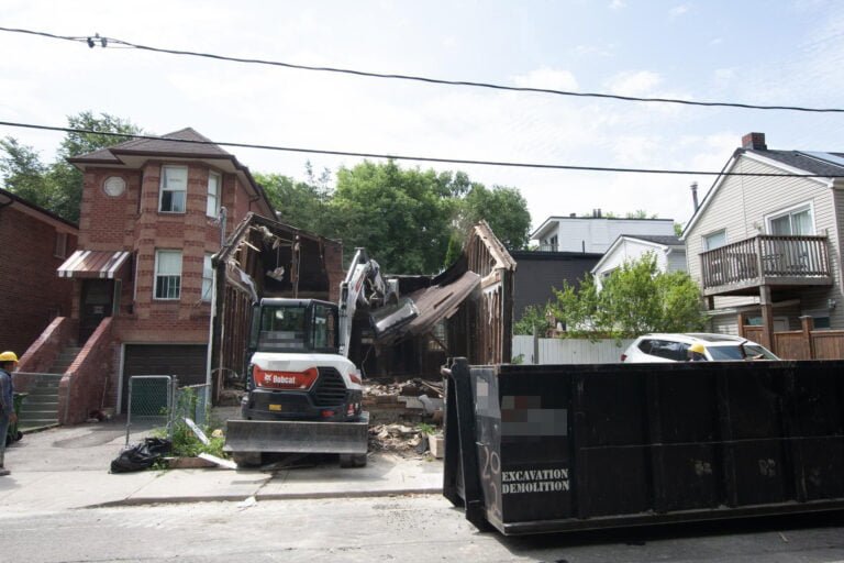 house demolition of a roof with an excavator, whilst two workers watch to assist where needed